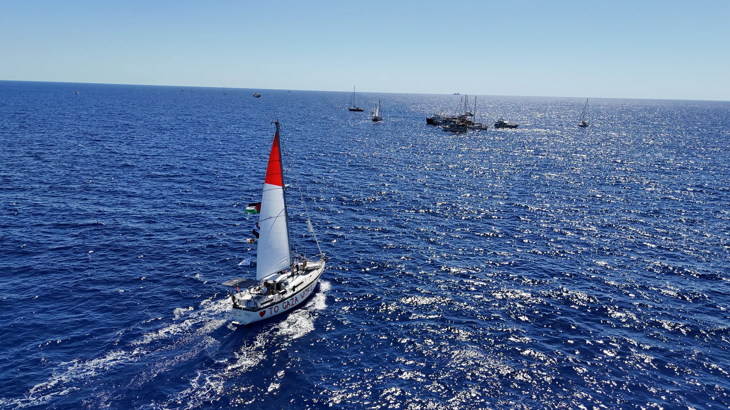 Aerial view of a sailboat with a red sail and Palestinian flag, hull painted 'To Gaza With Love,' leading a flotilla of boats across open water.